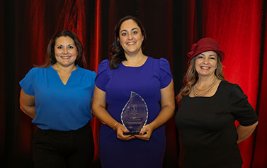 The 2024 Capstone Award Winner stands in between two colleagues in front of a red curtain while holding the curved, candle flame-shaped, glass award.