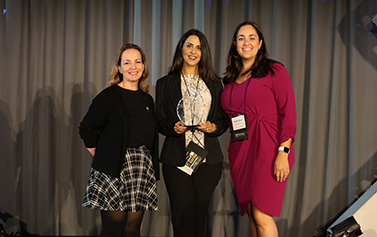 The 2025 Capstone Award Winner stands in between two colleagues in front of a gray curtain while holding the curved, candle flame-shaped, glass award.